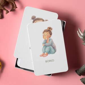 May include: A set of white emotion cards with a drawing of a girl labeled "BORED". The girl is sitting with her eyes closed. The cards are in a black box, with toy animals on a pink background.