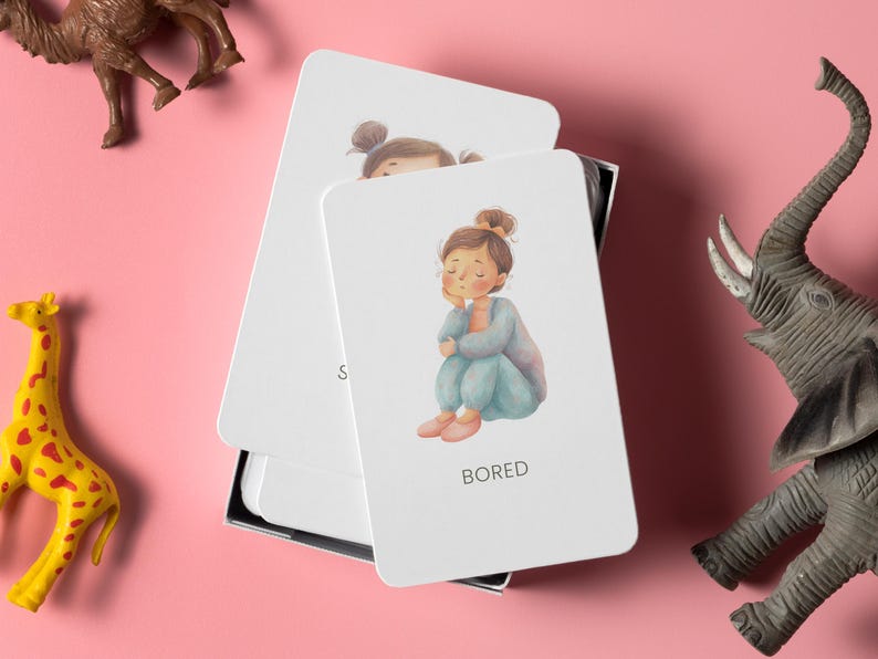 May include: A set of white emotion cards with a drawing of a girl labeled "BORED". The girl is sitting with her eyes closed. The cards are in a black box, with toy animals on a pink background.