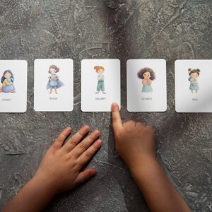 May include: Five emotion flashcards with illustrations and text: Lonely, Brave, Grumpy, Scared, and Sad. A child's hand points to the Grumpy card. The cards are on a textured gray surface.