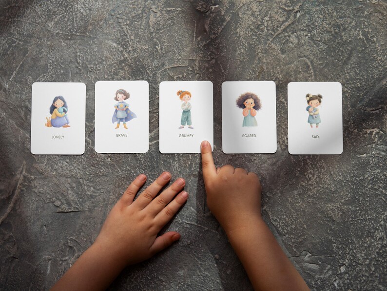 May include: Five emotion flashcards with illustrations and text: Lonely, Brave, Grumpy, Scared, and Sad. A child's hand points to the Grumpy card. The cards are on a textured gray surface.