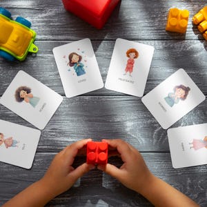 May include: A collection of emotion flashcards with illustrations of children expressing feelings like loved, frustrated, and happy. A child's hands hold a red building block, surrounded by toys on a gray wooden surface.