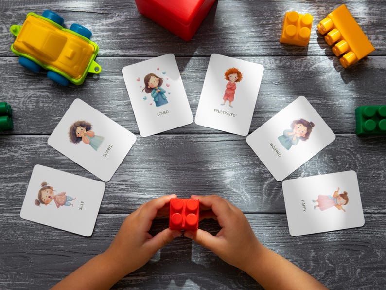 May include: A collection of emotion flashcards with illustrations of children expressing feelings like loved, frustrated, and happy. A child's hands hold a red building block, surrounded by toys on a gray wooden surface.