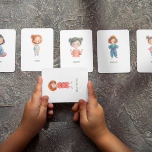 May include: A set of emotion flashcards with illustrations of children and corresponding emotion words: Calm, Tired, Angry, Proud, Silly, and Frustrated. The Frustrated card is held by a child's hands. The cards are on a gray surface.