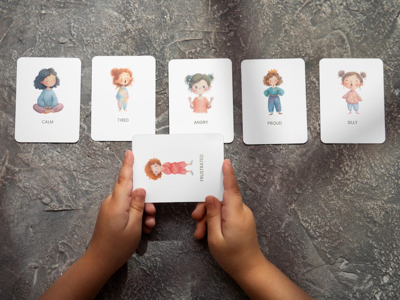 May include: A set of emotion flashcards with illustrations of children and corresponding emotion words: Calm, Tired, Angry, Proud, Silly, and Frustrated. The Frustrated card is held by a child's hands. The cards are on a gray surface.