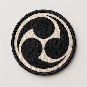 May include: A circular embroidered patch featuring a black triple tomoe symbol centered on a cream-colored background. The design is enclosed by a thick, black stitched border, creating a clean, high-contrast graphic appearance.