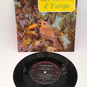 Könnte beinhalten: Eine Vintage-Schallplatte und ein Buch mit dem Titel "Les oiseaux d'Europe" werden gezeigt. Das Buchcover zeigt ein lebendiges Bild eines Vogels, der auf einem Ast sitzt. Die Schallplatte ist schwarz mit Text und Etiketten.