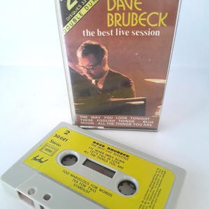 Può includere: Una cassetta e custodia vintage con "Dave Brubeck: The Best Live Session". La cassetta è grigio chiaro con un'etichetta gialla, che mostra i titoli delle canzoni. La custodia ha una copertina marrone con il nome dell'artista e il titolo dell'album.