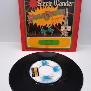Könnte beinhalten: Eine Vintage-Vinyl-Schallplatte und das Albumcover für Stevie Wonders "Master Blaster (Jammin')". Das Albumcover ist rot mit einem grünen Rand und zeigt den Titel in Gelb und den Namen des Künstlers. Die Schallplatte ist schwarz mit einem weiß-blauen Label.