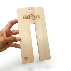 May include: A light-colored wooden display card with a rectangular cutout in the center and a small hole at the top. The text "fátia's kraftshop" is engraved near the top, and a small heart icon is engraved near the bottom left corner.