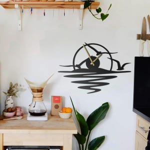 May include: Black metal wall clock featuring a sunset design with seagulls in flight. The clock has gold-colored hands and is mounted on a white wall. The clock's design includes a circular sun and water reflections.