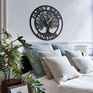 Könnte beinhalten: Schwarze Metall-Wanduhr mit einem Lebensbaum-Design. Das Zifferblatt zeigt die Worte "FAMILY NAME CUSTOM" in kreisförmiger Anordnung. Goldfarbene Zeiger zeigen die Uhrzeit an. Die Uhr ist an einer weißen Wand über einem Bett mit Kissen befestigt.