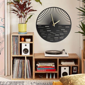 May include: A black and gold wall clock with a sunburst design hangs above a wooden record player stand. The stand holds a white turntable, speakers, and stacks of vinyl records. A potted plant sits on top of a wooden shelf unit.