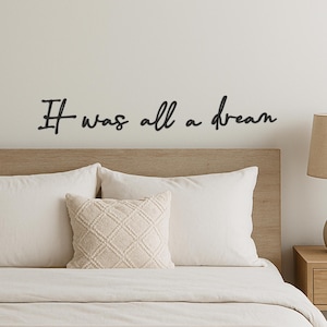 Puede incluir: Una escena de dormitorio con una cama con ropa de cama y almohadas blancas. Una almohada decorativa con un patrón de diamantes se encuentra frente a las almohadas blancas. Encima de la cama, la frase "It was all a dream" está escrita en guión negro.