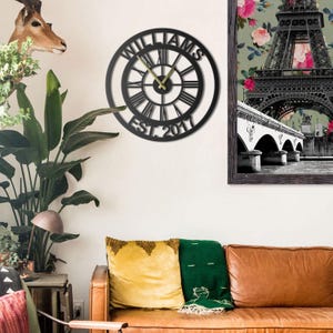 Può includere: Orologio da parete in metallo nero con numeri romani e il nome "WILLIAMS" sul bordo esterno. Le lancette dell'orologio sono dorate. L'anno "EST. 2017" è visualizzato in basso. L'orologio è appeso a una parete bianca.