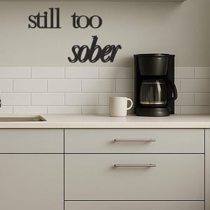 May include: Black metal wall art spelling "still too sober" above a kitchen counter. A black coffee maker and a white mug are on the counter. The wall is light gray with white subway tile.