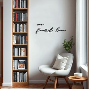 Può includere: Una libreria in legno piena di libri è accanto a un muro bianco con la frase "we found love" in corsivo nero. Sono visibili anche una sedia bianca con un cuscino e un tavolino con una ciotola.