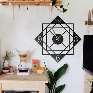 Op de afbeelding: Een zwarte geometrische wandklok met gouden wijzers en de cijfers 12, 3, 6 en 9. De klok is aan een witte muur bevestigd. Het ontwerp van de klok is een vierkant in een ruit. De klok maakt deel uit van een keukeninrichting.