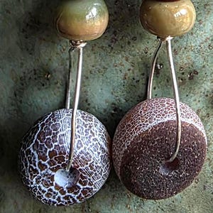Può includere: Un paio di orecchini pendenti con ganci in argento, piccole perline sfaccettate rosso scuro, perline lisce verde oliva e perline circolari più grandi con una superficie testurizzata e screpolata nei toni del marrone, bianco e grigio.