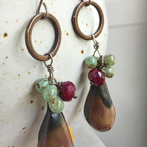 May include: A pair of dangle earrings featuring copper-toned metal hoops and wire-wrapped clusters of small, faceted mint green and deep red beads. Each earring is finished with a smooth, teardrop-shaped brown and black ceramic pendant.