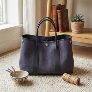 May include: A dark purple canvas tote bag with black leather trim, two top handles, and a central silver-toned snap button closure. The bag features a structured rectangular base and reinforced leather vertical straps extending from the handles.