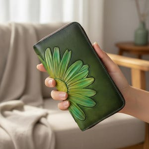 May include: Green leather wallet featuring an embossed daisy design. The wallet has a zip closure and a dark green border. The daisy is a gradient of green and yellow. The wallet is held in a hand.