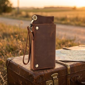 May include: A dark brown leather wallet featuring a vertical rectangular shape, a side zipper closure, a matching leather wrist strap, and two metallic snap buttons on the front flap. The wallet has a smooth, matte finish and a metal ring attachment.
