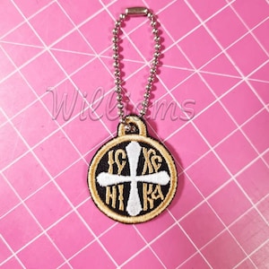 May include: Embroidered pendant with a cross design, featuring gold, black, and white colors. The pendant is attached to a silver ball chain. The text "IC XC NI KA" is visible on the pendant. The background is a pink cutting mat.