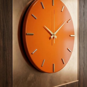 Puede incluir: Un reloj de pared circular con una esfera de color naranja vibrante y marcadores de hora dorados finos. El reloj cuenta con manecillas doradas delgadas para las horas, minutos y segundos. Está montado sobre una placa cuadrada de color cobre.