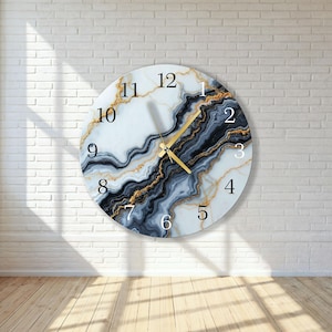 Puede incluir: Reloj de pared redondo con diseño de mármol blanco con vetas negras, doradas y grises. La esfera del reloj tiene números negros y manecillas doradas. El reloj está montado en una pared de ladrillo blanco.
