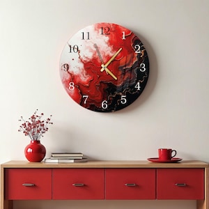 Puede incluir: Un reloj de pared redondo con un diseño abstracto rojo, negro y blanco. La esfera del reloj presenta manecillas doradas y números negros. Está montado en una pared blanca sobre un mueble rojo, un jarrón rojo y una taza y platillo rojos.