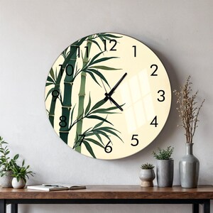 May include: A round wall clock with a cream-colored face featuring a green bamboo stalk illustration. The clock has black Arabic numerals from 1 to 12 and thin black hour, minute, and second hands. The design is printed on a smooth, reflective surface.