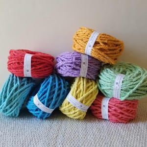 May include: A collection of colorful yarn skeins in shades of red, blue, yellow, green, purple, and orange. Each skein is wrapped with a white label. The yarn is stacked on top of each other.