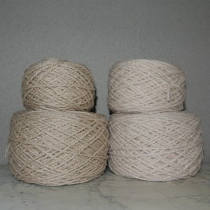 Puede incluir: Cuatro madejas de hilo beige apiladas en dos pares. El hilo está bien enrollado, mostrando la textura de las fibras. El color neutro y la fibra natural sugieren que el hilo es adecuado para proyectos de tejido o ganchillo.