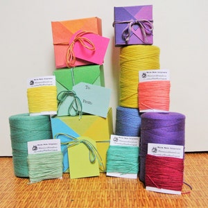 May include: A collection of colorful gift boxes and spools of twine. The boxes are in shades of orange, green, purple, and yellow, tied with matching twine. Several spools of twine are also visible, in various colors, with small white labels.