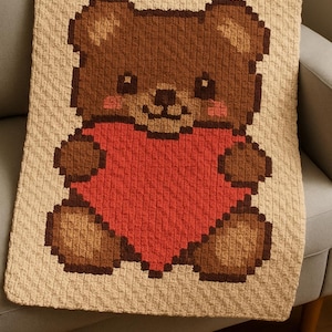 Könnte beinhalten: Handgefertigte Häkeldecke mit einem pixeligen Teddybären, der ein rotes Herz hält. Der Bär ist braun mit beigen Akzenten und einem lächelnden Gesicht. Die Decke ist beige und liegt auf einem grauen Sofa.
