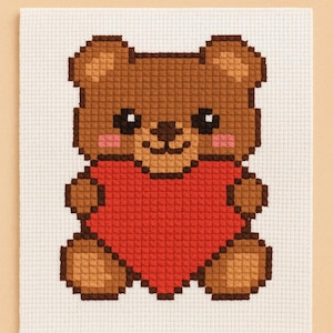 Op de afbeelding: Kruissteekontwerp van een bruine teddybeer met een rood hart. De beer heeft een lachend gezicht met zwarte ogen en roze wangen. Het ontwerp staat op een witte stoffen achtergrond.