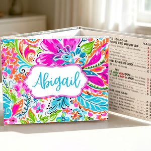 Può includere: Un portamenu colorato con un motivo floreale rosa, blu, arancione e verde. Il nome "Abigail" è scritto in blu chiaro all'interno di una cornice bianca. Il portamenu è aperto, rivelando un elenco di articoli e prezzi.