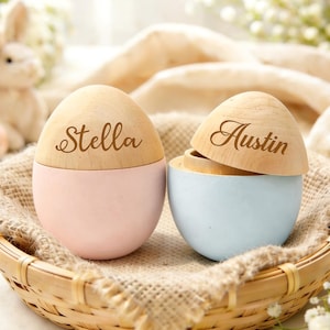 May include: Two wooden egg-shaped boxes, one pink and one blue, sit in a woven basket. The pink egg is labeled "Stella," and the blue egg is labeled "Austin." The lids are light wood, and the bases are painted.