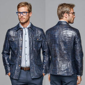 Pode incluir: Casaco de couro azul marinho com textura de pele de crocodilo. O casaco tem quatro bolsos frontais, botões dourados e um cinto castanho. O modelo usa uma camisa azul clara e óculos. Peça de roupa exterior elegante.