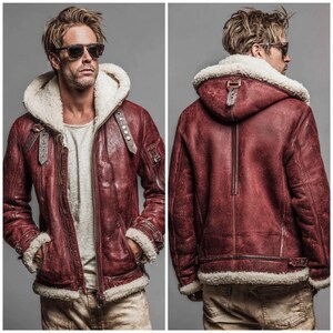 May include: A burgundy leather jacket with a cream-colored shearling lining and hood. The jacket features a zip-up front, multiple pockets, and buckle details. The model is wearing sunglasses and a light-colored shirt. The jacket is a winter outerwear item.