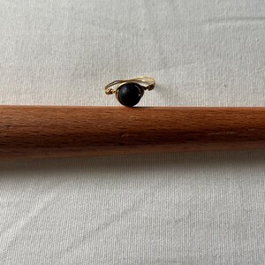 Peut inclure: Une bague faite à la main avec une bande de fil de couleur or. La bague présente une pierre sombre et ronde au centre, enveloppée solidement par le fil. La bague est présentée sur une cheville en bois sur un fond de tissu blanc.