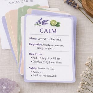 Könnte beinhalten: Ein Stapel "CALM CARDS" für ätherische Öle mit einer Mischung aus Lavendel und Bergamotte. Die Karten sind weiß mit lila und grünen Akzenten und enthalten Anweisungen. Getrockneter Lavendel und eine Holzschale befinden sich im Hintergrund.