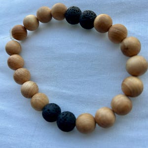 May include: A beaded bracelet featuring light brown wooden beads and black lava stone beads. The bracelet is strung on a clear elastic cord. The beads are round and smooth, creating a natural and earthy aesthetic.