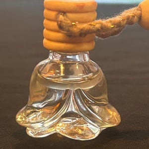 May include: A clear glass car air freshener with a flower-shaped bottom and a wooden top. The wooden top is made of stacked rings and is connected to a rope for hanging. The liquid inside is light amber.