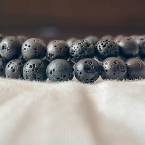 May include: Close-up of two black lava stone bead bracelets. The beads are round and have a porous texture. The bracelets are resting on a white, textured surface. The bracelets are a dark gray color.