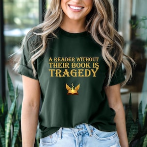 Peut inclure: T-shirt vert foncé avec l'inscription dorée « A READER WITHOUT THEIR BOOK IS TRAGEDY ». En dessous, un dessin de livre ailé. Le t-shirt est fait d'une matière douce.
