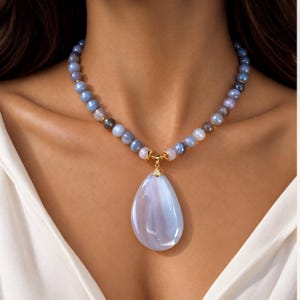 Puede incluir: Un collar con una hilera de cuentas redondas de color azul claro y gris, con un colgante grande, liso y en forma de lágrima de color azul claro. El colgante está unido a la hilera de cuentas mediante un engaste de metal dorado con una piedra.