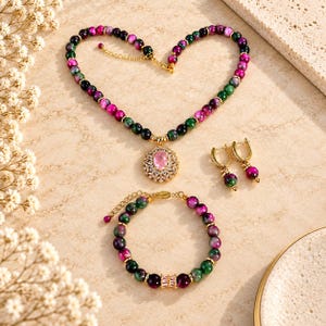 Puede incluir: Un conjunto de joyería que consta de un collar, una pulsera y un par de pendientes de aro. El collar y la pulsera presentan cuentas alternas de color verde oscuro y magenta con detalles dorados. El collar incluye un colgante ovalado rosa.