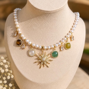 Puede incluir: Un collar de perlas con detalles dorados y varios colgantes de piedras preciosas. El collar presenta un colgante de sol y piedras cuadradas y rectangulares en tonos verdes, marrones y blancos. El collar se exhibe en un maniquí beige.
