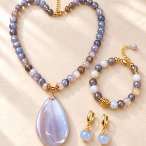 Puede incluir: Un conjunto de joyería que consta de un collar, una pulsera y un par de pendientes. El collar presenta cuentas facetadas grises y blancas con un gran colgante liso en forma de lágrima. La pulsera tiene cuentas a juego y un espaciador de filigrana dorada. Los pendientes son aros dorados con cuentas azul claro colgantes.